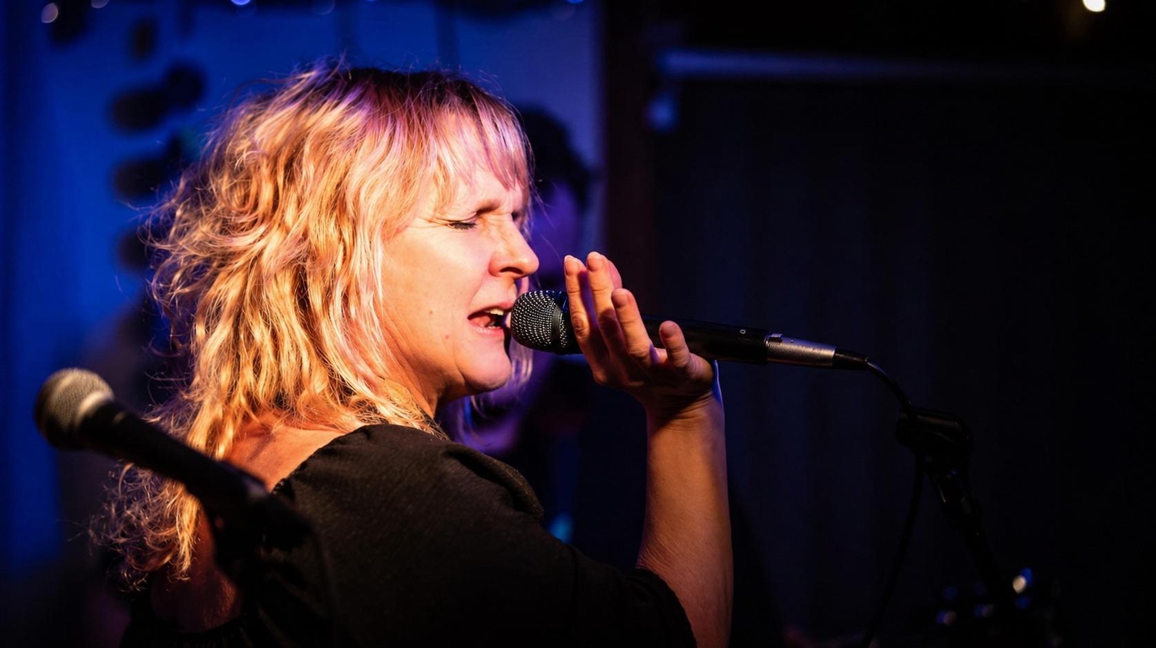Tonya Horoky Cooper performing live, eyes closed, microphone in hand, lit by warm and blue stage light